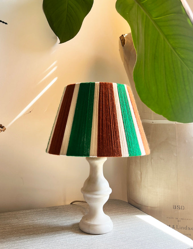 A table lamp with a white pedestal base and Slice Shades yarn lampshade shade featuring vertical stripes of green, brown, cream, and orange, styled on a tabletop with warm sunlight and large green leaves in the background.