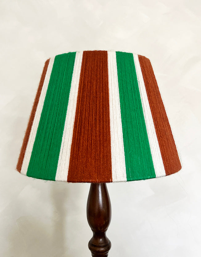 A table lamp with a Slice Shades yarn lampshade featuring vertical stripes of green, rust brown, and white, mounted on a dark brown turned wood base against a light textured wall.
