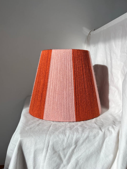 A Slice Shades handwoven yarn striped lampshade with vertical stripes in rust orange and light pink, photographed against a wrinkled white fabric backdrop.