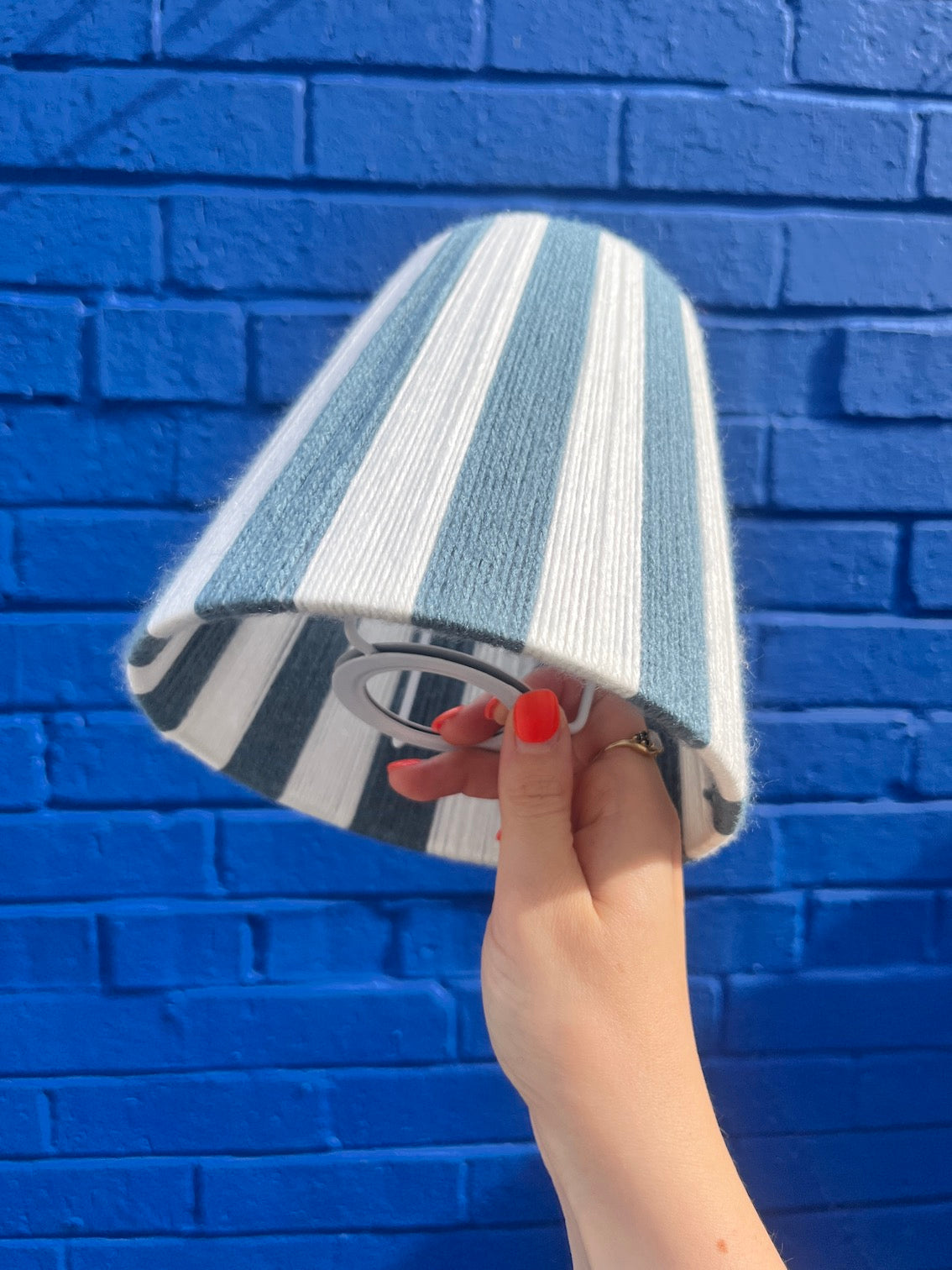 A hand holds a blue and white striped yarn Slice Shades lampshade in front of a bright blue brick wall.