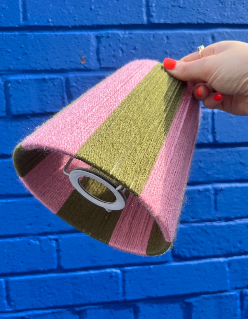 A hand holding a Slice Shades yarn lampshade with vertical stripes of pink, and olive green set against a bright blue painted brick wall.