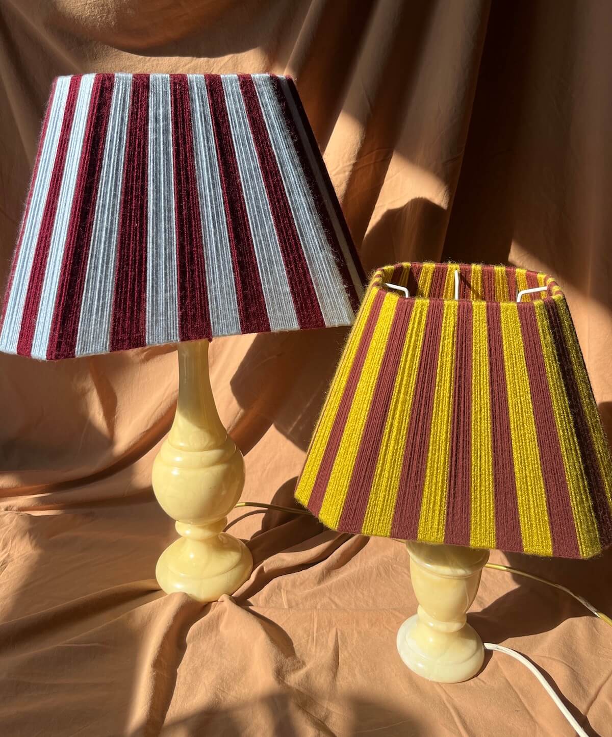 Two Sice Shades table lamps with cream-colored turned bases and striped yarn shades featuring vertical stripes in burgundy and light blue, and the other in chartreuse and burgundy, set against a draped tan fabric background.