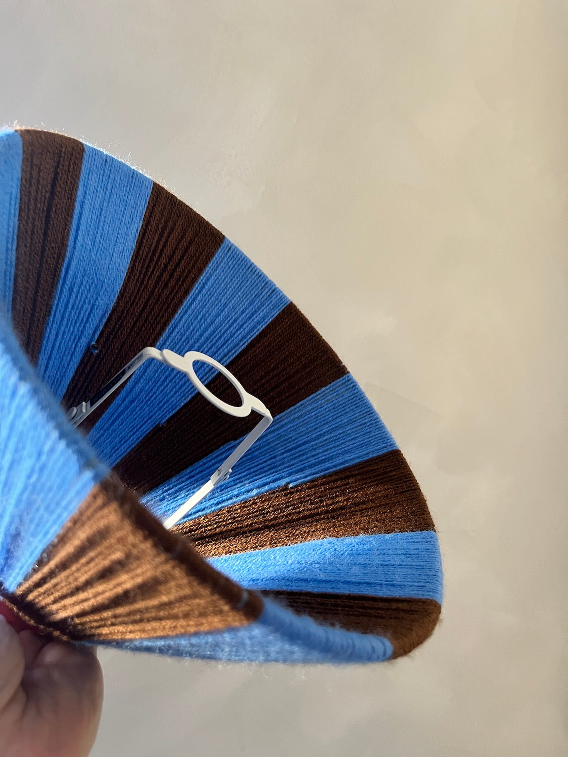 A close-up of a hand holding a Slice Shades lampshade made of alternating blue and brown striped yarn, with a white plastic wire frame visible inside.