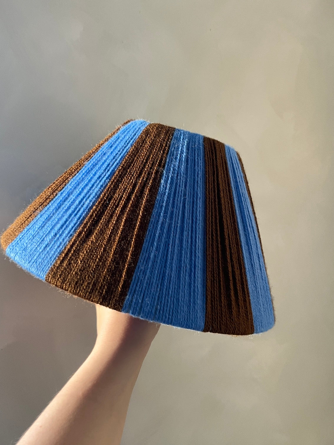 A Slice Shades yarn lampshade with alternating vertical stripes of medium blue and dark brown, held up by a person's hand against a plain light gray background.