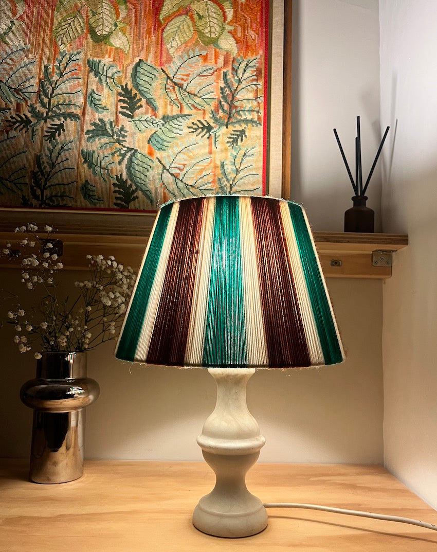 A table lamp with a striped Slice Shades yarn lampshade in green, brown, cream, and white, mounted on a cream-coloured ceramic base, set on a wooden table in a decorated room.