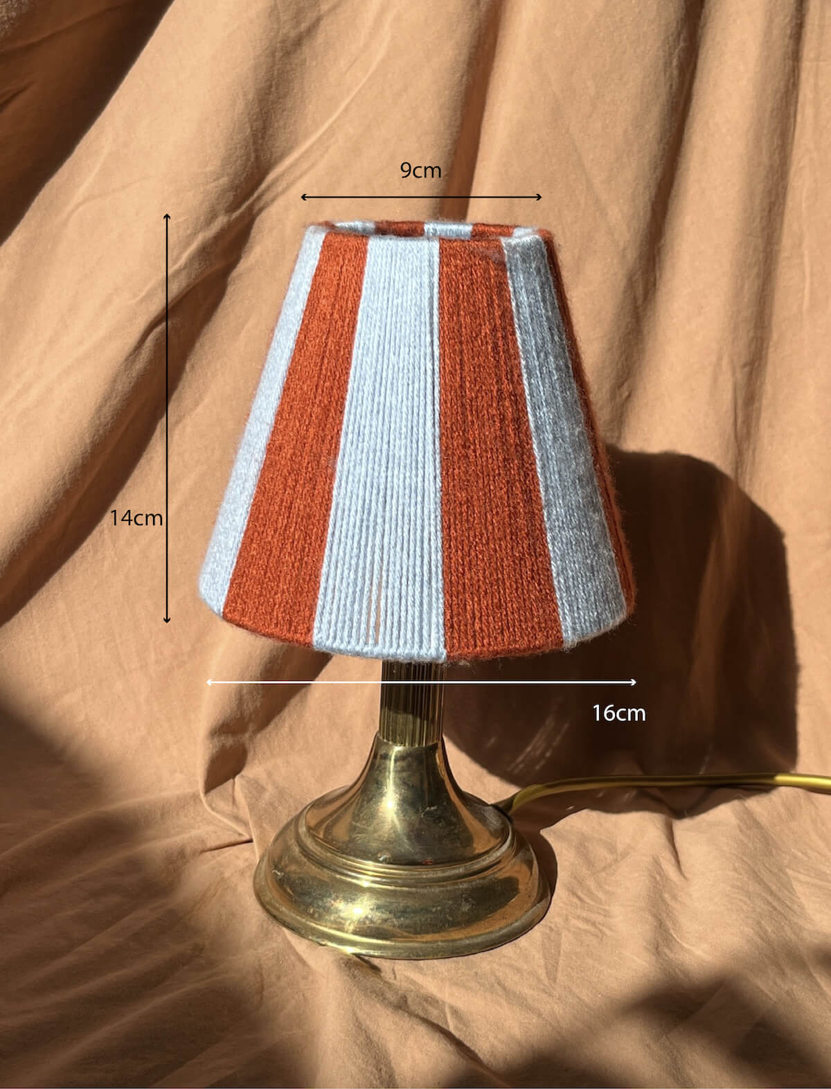 A Slice Shades handmade string yarn lampshade with alternating rust orange and light blue stripes, photographed on a draped tan fabric background.