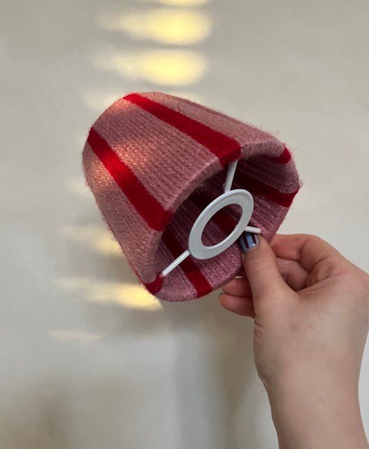 A hand holding a Slice Shades yarn lampshade in a dusty pink and red striped pattern, with a white plastic ring visible on the side, against a softly lit off-white background.